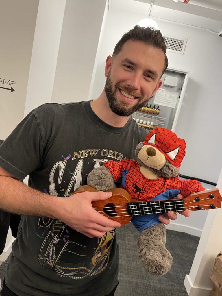 A smiling man holds a teddy bear dressed in a Spider-Man costume while also holding a small ukulele. The man is participating in a Toys for Tykes team building event, proudly showcasing the decorated toy ready to be donated to a child.