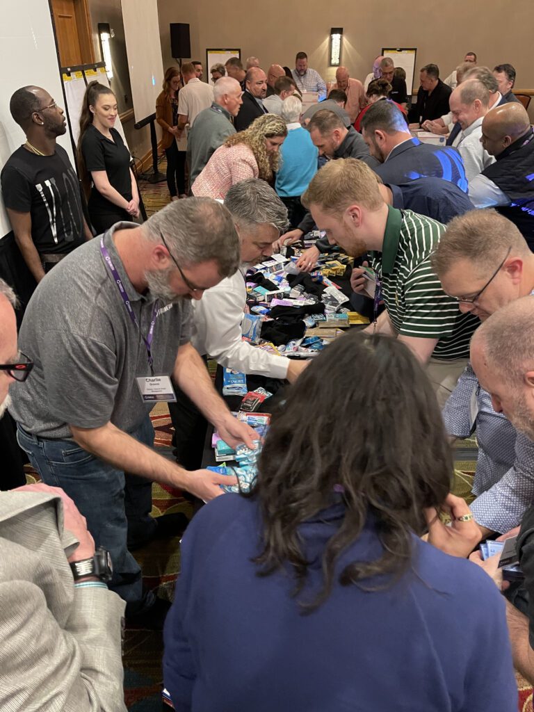 Teams gather around a table, enthusiastically packing military care packages during a team building event to support troops.