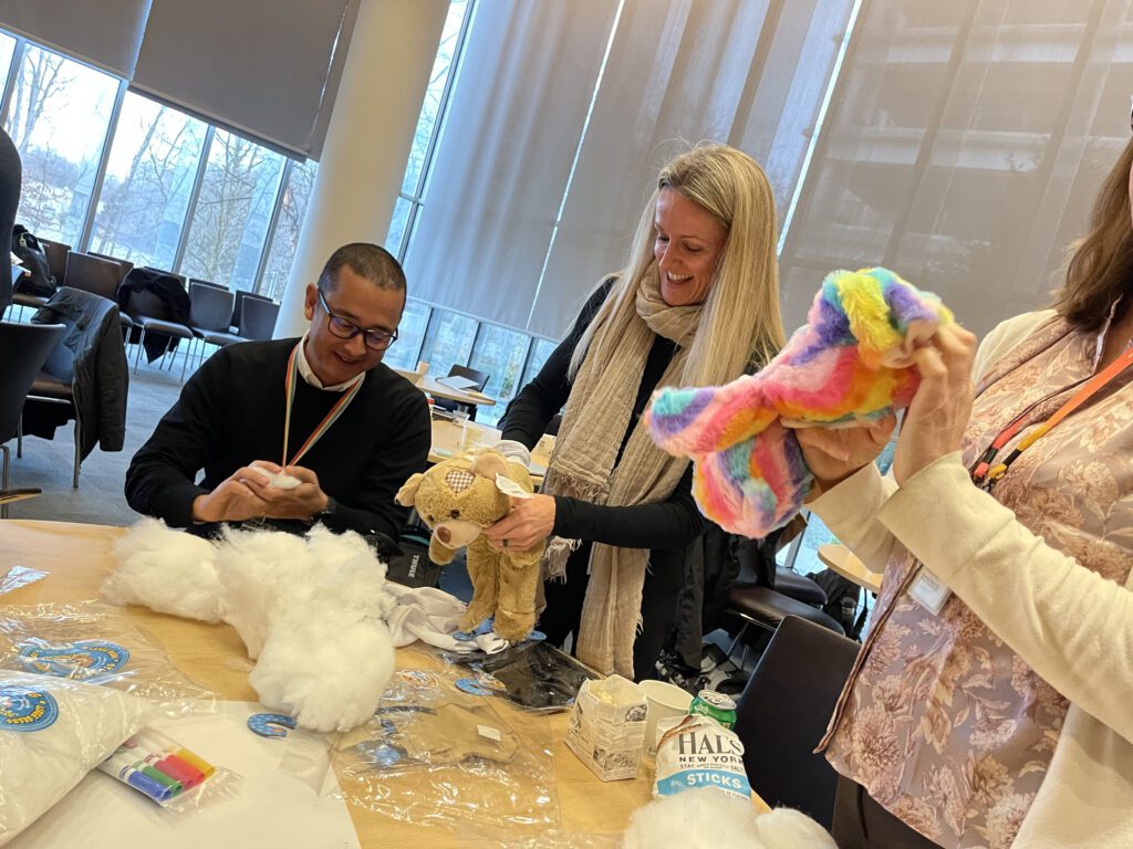 Three participants in the Team Teddy Rescue Bear team building activity are gathered around a table filled with teddy bear stuffing and various craft supplies. A woman is holding a colorful rainbow-patterned teddy bear, while a man next to her is assembling another bear.