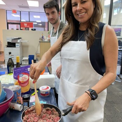 A woman and a man participate in a culinary team building event. The woman is smiling as she stirs a pan filled with ingredients while wearing a white apron. Various food items like cheese and sour cream are visible on the table in front of them. The man behind her is also wearing an apron, standing by the food preparation area.