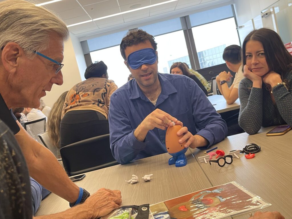 A group of people participates in a Toys for Tykes team building activity. One man is blindfolded while assembling a Mr. Potato Head toy, with other team members observing and giving instructions, creating a fun and collaborative atmosphere.