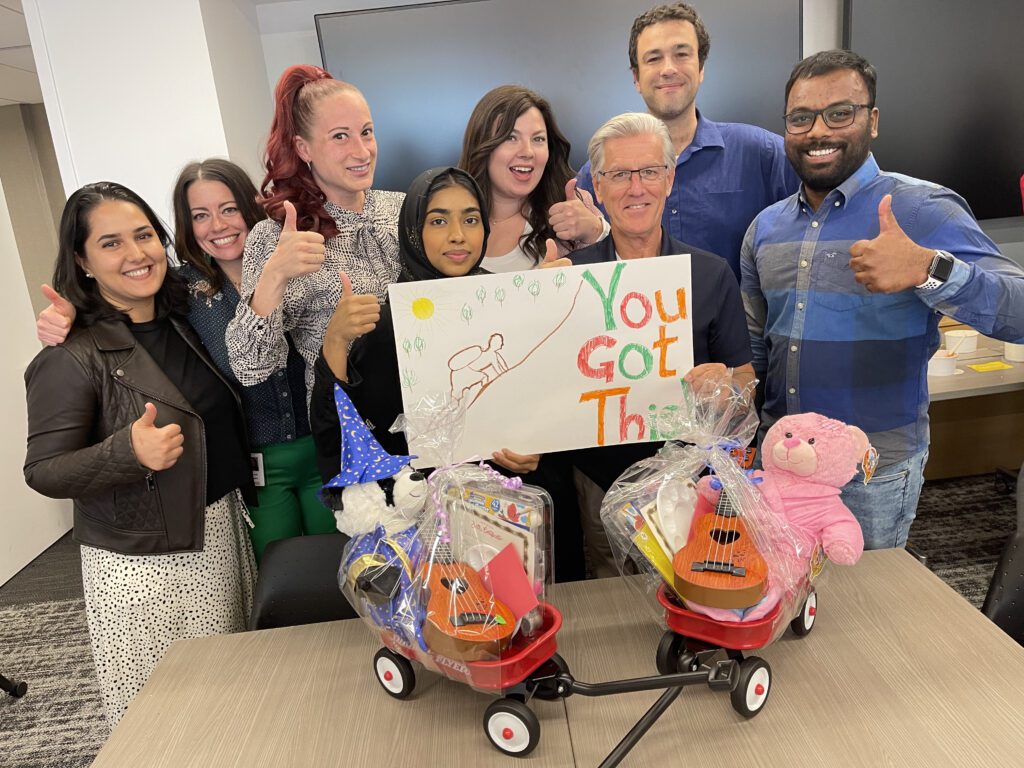 A group of eight people stand together smiling and giving thumbs up around two small Radio Flyer wagons filled with teddy bears and toys. One person is holding a handmade sign that says 'You Got This!' with a colorful drawing. The team appears happy and proud, showcasing their contributions during a team building activity focused on charity and giving back.