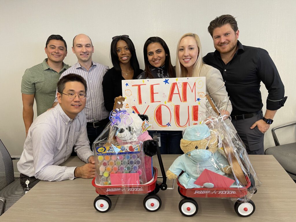 A team of six people proudly showcases their completed toy wagons filled with stuffed animals, arts and crafts supplies, and a sign that reads 'TEAM YOU!' as part of a Toys for Tykes team building event.