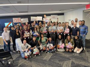 A large group of people smiling and posing together in a modern office space. They are holding and sitting with small red Radio Flyer wagons filled with various toys, teddy bears, and musical instruments, all wrapped in gift wrap. Behind them, some participants are holding up hand-drawn signs with motivational messages like 'You Got This!' This photo captures the conclusion of the Toys for Tykes ‘Arts & Music’ CSR team building event.