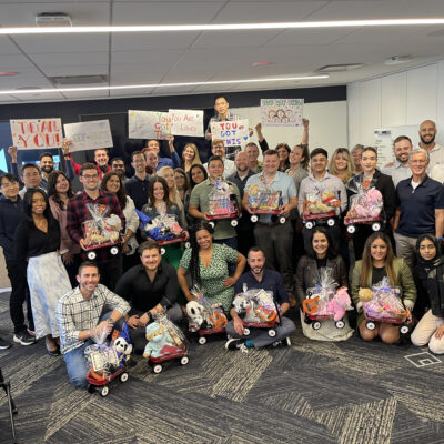 A large group of people smiling and posing together in a modern office space. They are holding and sitting with small red Radio Flyer wagons filled with various toys, teddy bears, and musical instruments, all wrapped in gift wrap. Behind them, some participants are holding up hand-drawn signs with motivational messages like 'You Got This!' This photo captures the conclusion of the Toys for Tykes ‘Arts & Music’ CSR team building event.