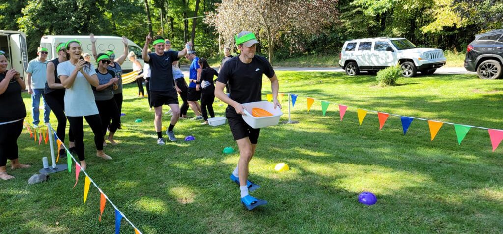 Participant balancing a tray with items while walking through an obstacle course as teammates cheer during the Outrageous Games team building event.