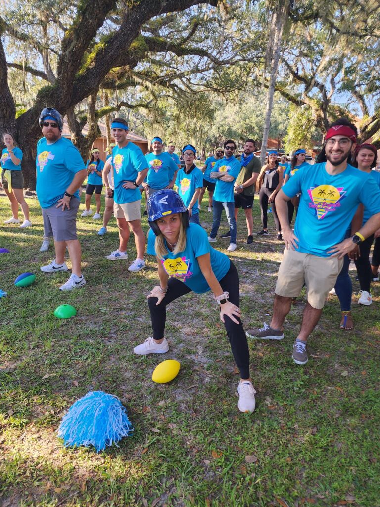 Team members lined up and ready for an outdoor relay race during the Outrageous Games team building event, showcasing their team spirit in matching blue shirts.