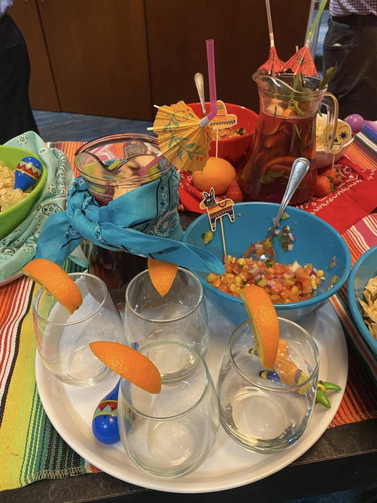 A festive spread featuring glasses garnished with orange slices, a pitcher of colorful drink, fresh salsa, and bowls of chips, all part of the Salsa Showdown team building event.