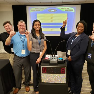 A team of five participants posing with enthusiasm and pointing to the screen, showing a game show setup during the Survey Says team building activity, highlighting team spirit, competition, and fun.