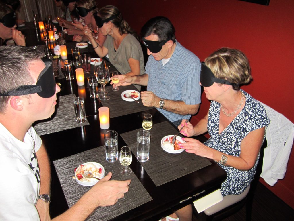 Participants enjoying a meal in a Dining in the Dark experience, with blindfolds on as they engage their senses of taste and smell while sharing the experience.