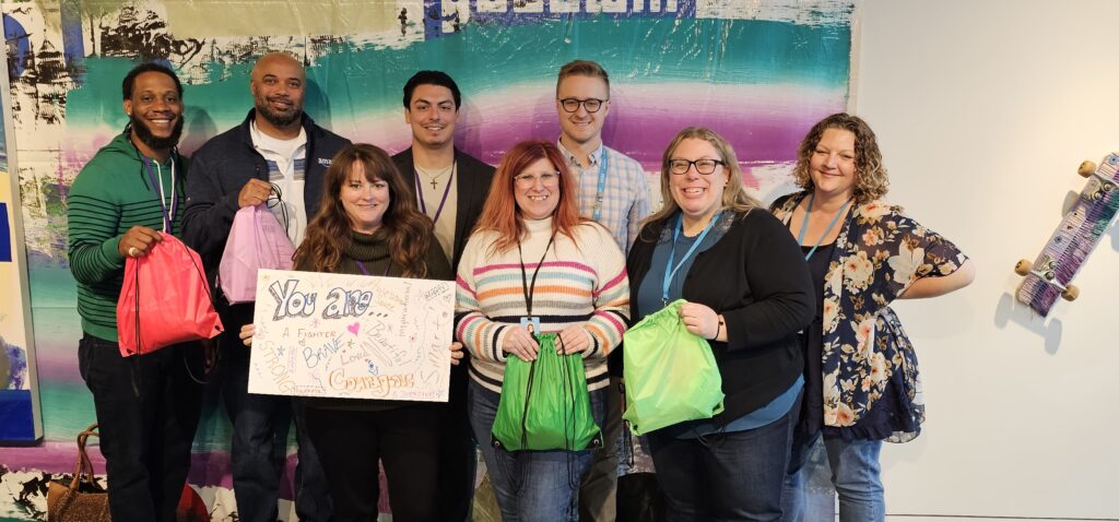 A group of participants smiles while holding colorful drawstring bags filled with care kits during a customized team building event based on the Helping Hands program. One participant holds a sign that reads 'You are amazing!' as they celebrate their contributions.