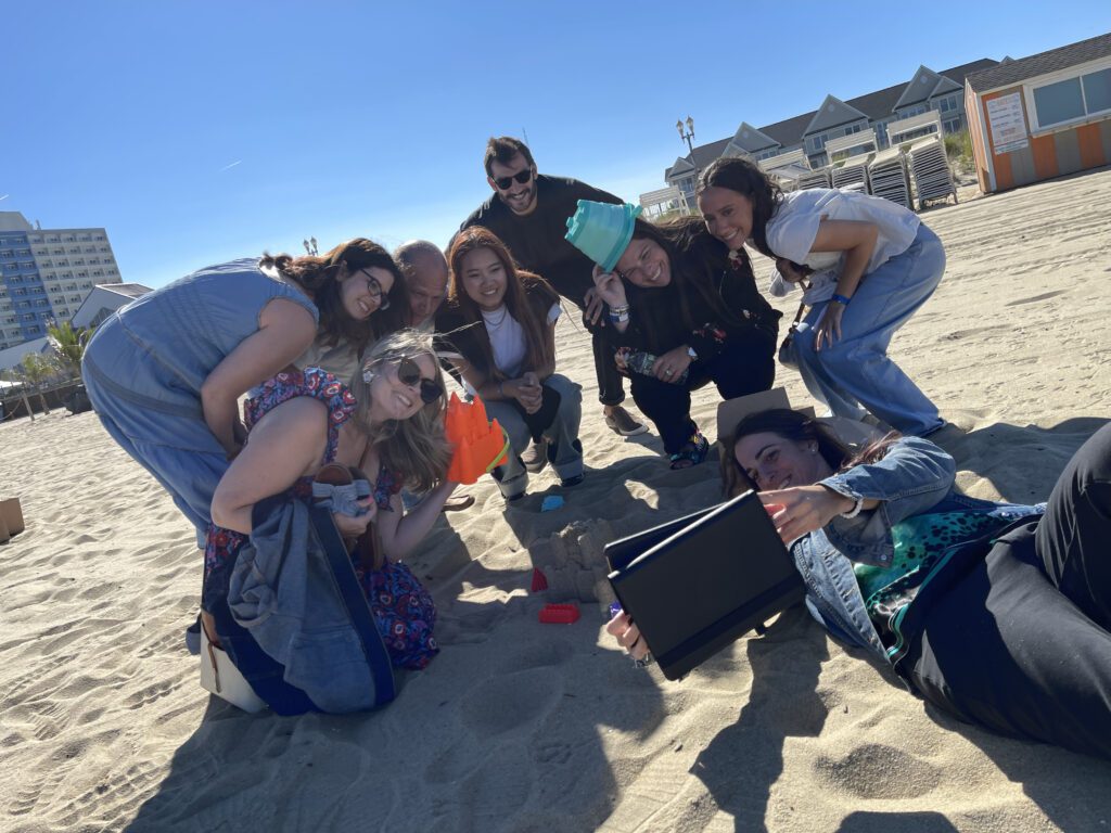 A group of participants poses together on the beach while building a sandcastle during a Custom Team Building event. They are smiling and using a tablet to capture the fun moment in the sun.