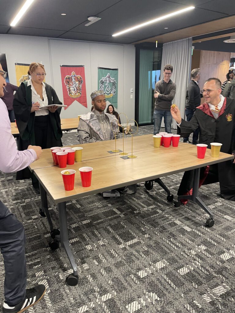 Participants dressed in wizard and knight costumes play a Harry Potter themed game involving cups and a ball during a Custom Team Building event. The room is decorated with banners representing different houses, adding to the magical atmosphere of the activity.