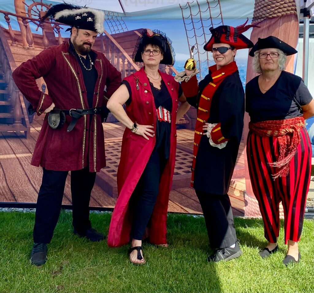 Four participants dressed as pirates pose confidently in front of a pirate ship backdrop during a Custom Team Building event. Their themed attire showcases the creativity and customization of the team building experience.