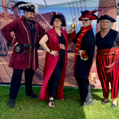 Four participants dressed as pirates pose confidently in front of a pirate ship backdrop during a Custom Team Building event. Their themed attire showcases the creativity and customization of the team building experience.