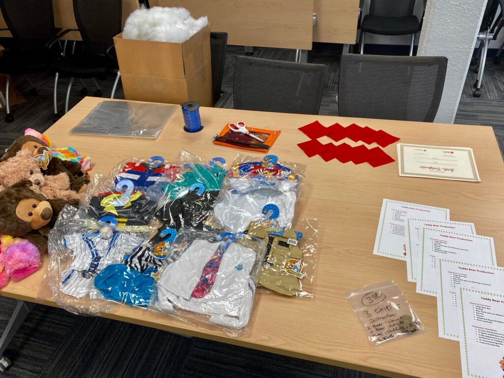 Table setup with teddy bear outfits, materials, and instructions for teams to create stuffed animals as part of The Big Give team building event, where all items will be donated to children in need.