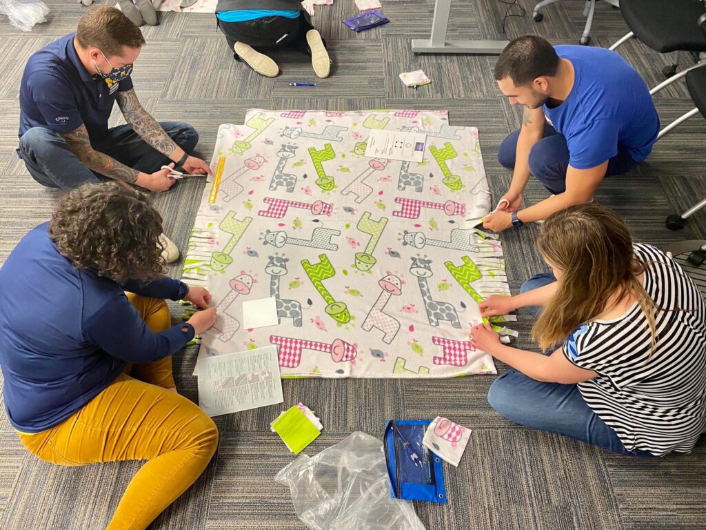 Participants work diligently on creating a fleece blanket with playful animal prints as part of The Big Give team building event, contributing to a charitable cause.