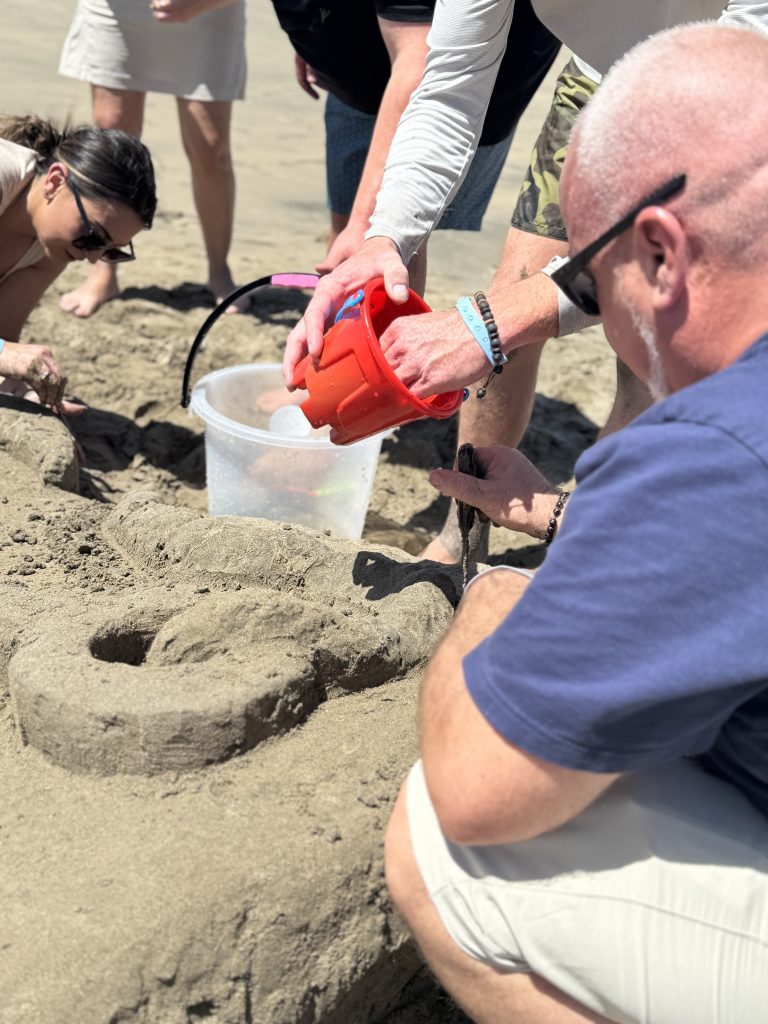 Team members collaborating on their sand sculpture, using buckets and tools to shape the design during a sand sculpting team building event at the beach.