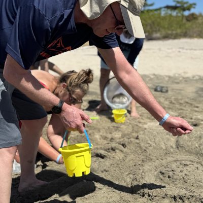 Participants working together at a sand sculpting team building event, using small buckets to gather sand for their sculpture on the beach.