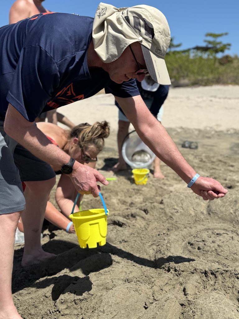 Participants working together at a sand sculpting team building event, using small buckets to gather sand for their sculpture on the beach.