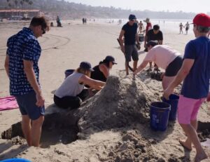 A team working together at the beach to build a large sand sculpture during a sand sculpting team building event, with teamwork and coordination.