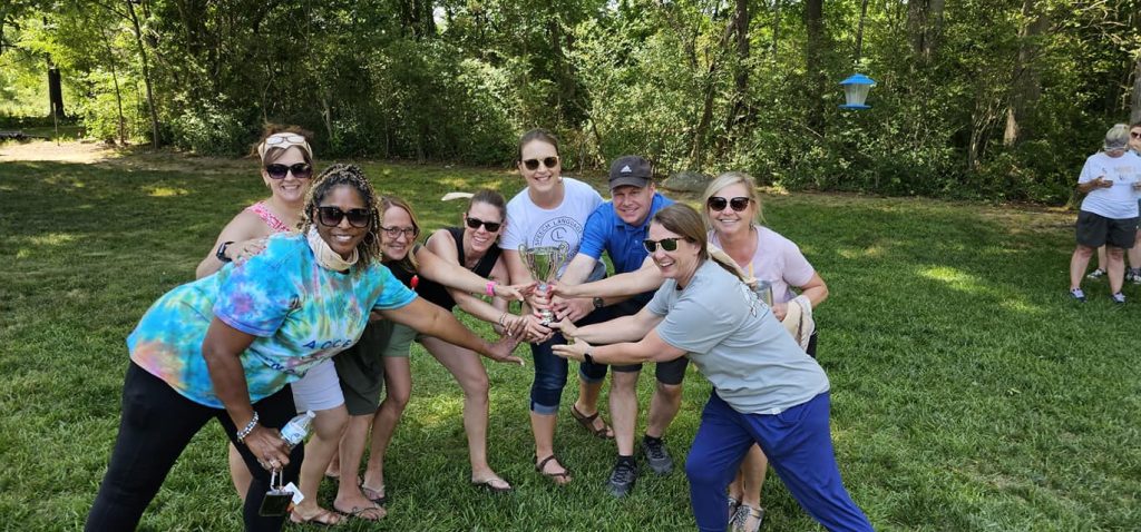 A group of teammates smiling and celebrating with a trophy at the Ultimate Tailgate Challenge, posing in the outdoors with classic tailgating games in the background.