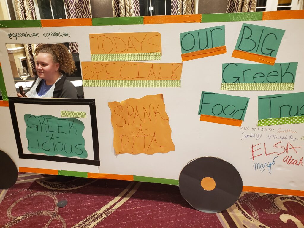 The image showcases a creative and colorful food truck facade crafted for a team building event, focusing on a Greek food theme. The food truck, labeled 