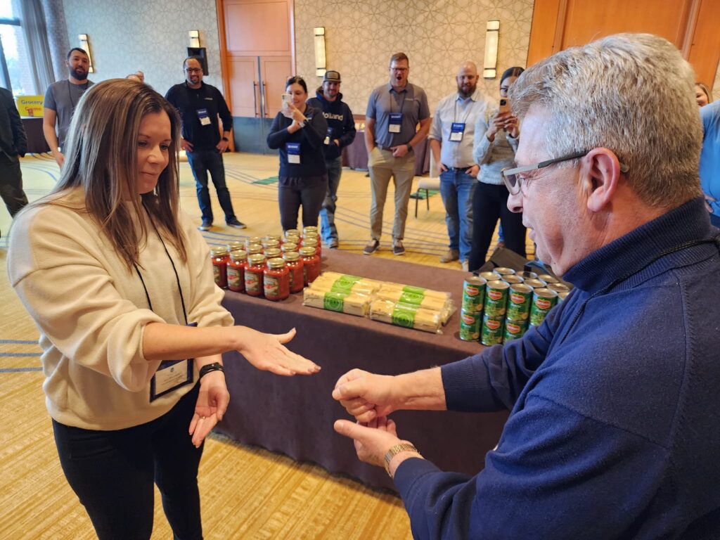 Two participants engage in a rock-paper-scissors game during the Foodbank Showdown team building event. A table filled with canned goods and pasta in the background showcases the charitable aspect of the event.