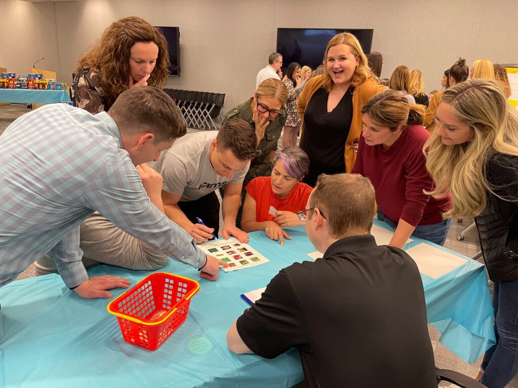 Team collaborates on a Foodbank Showdown challenge, using teamwork and strategy to complete a grocery-themed activity while preparing care packages for donation to local families in need.