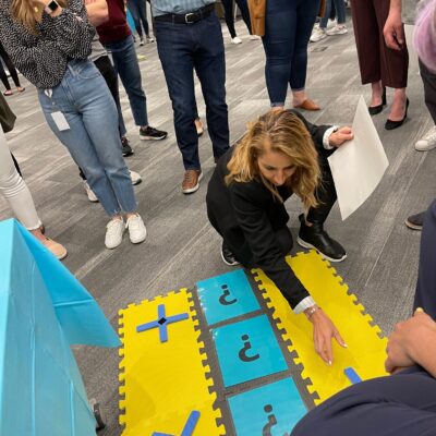 A participant carefully places a marker on a large tic-tac-toe style game board during a Foodbank Showdown team building event. The surrounding group watches intently as the competition unfolds.