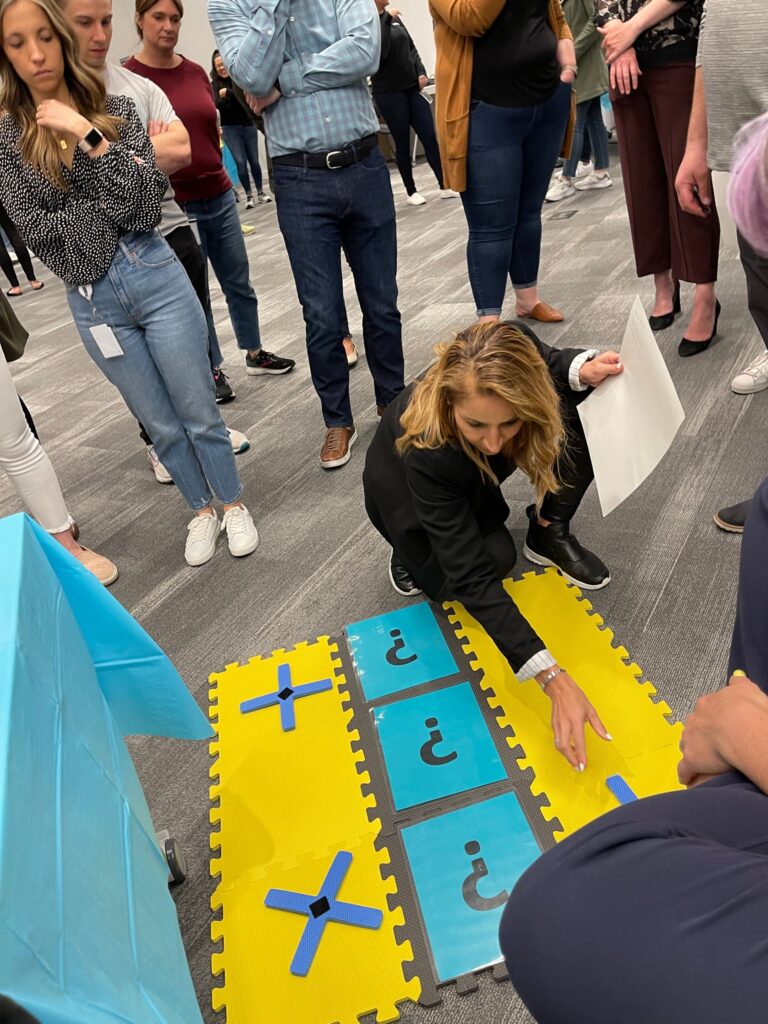 A participant carefully places a marker on a large tic-tac-toe style game board during a Foodbank Showdown team building event. The surrounding group watches intently as the competition unfolds.