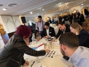 A group of participants collaborates at a table during the 'Foodbank Showdown' team building event. They review materials and work together to complete a grocery-themed challenge, contributing to the charitable initiative by earning points to purchase groceries for donation to local families in need.