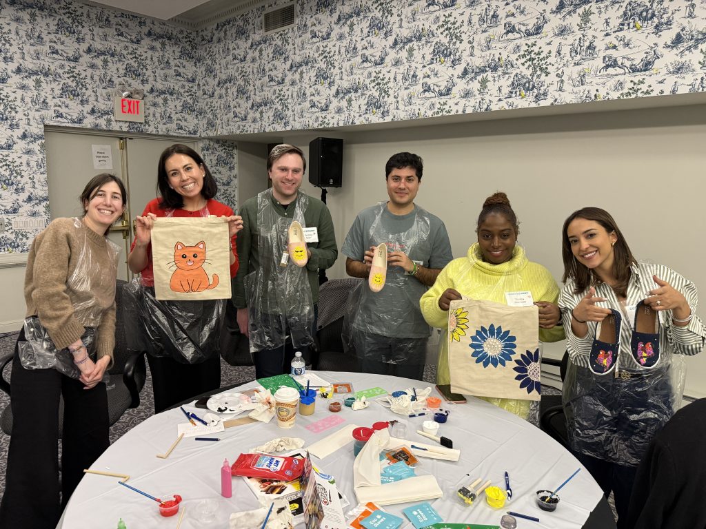 Participants proudly display their painted TOMS shoes and tote bags during the Paint Soles for Souls team building event, showcasing vibrant designs including a cat and a flower.