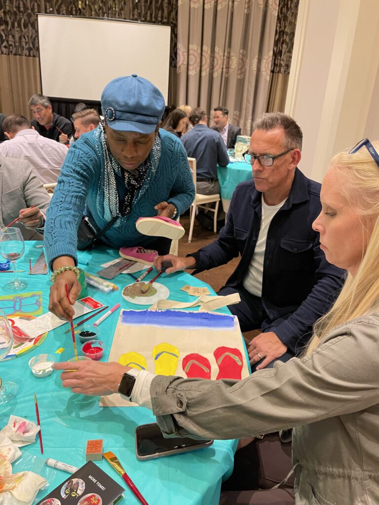 Participants work together to paint TOMS shoes and tote bags during the Paint Soles for Souls charitable team building event. One participant paints a tote bag with colorful flip-flop designs while others decorate shoes with intricate patterns.