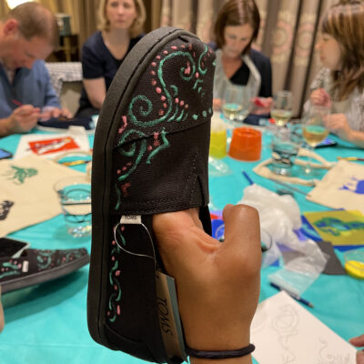 A close-up of a hand-painted TOMS shoe decorated with intricate, colorful designs during the Paint Soles for Souls charitable team building event. Participants in the background are focused on painting additional shoes and tote bags. The event combines creativity, teamwork, and philanthropy to make a positive global impact.