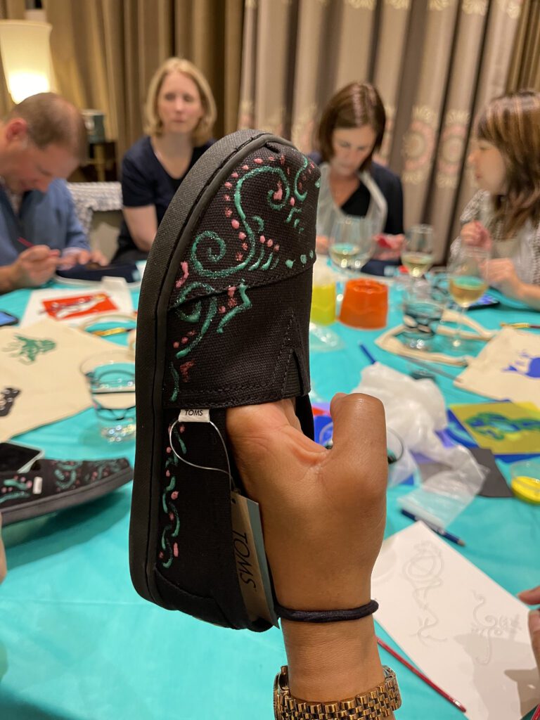 A close-up of a hand-painted TOMS shoe decorated with intricate, colorful designs during the Paint Soles for Souls charitable team building event. Participants in the background are focused on painting additional shoes and tote bags. The event combines creativity, teamwork, and philanthropy to make a positive global impact.