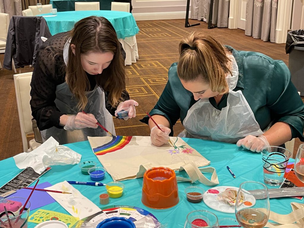 Two women wearing aprons are focused on painting tote bags with colorful designs, including rainbows and flowers, during the Paint Soles for Souls charitable team building event.