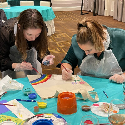 Two women wearing aprons are focused on painting tote bags with colorful designs, including rainbows and flowers, during the Paint Soles for Souls charitable team building event.