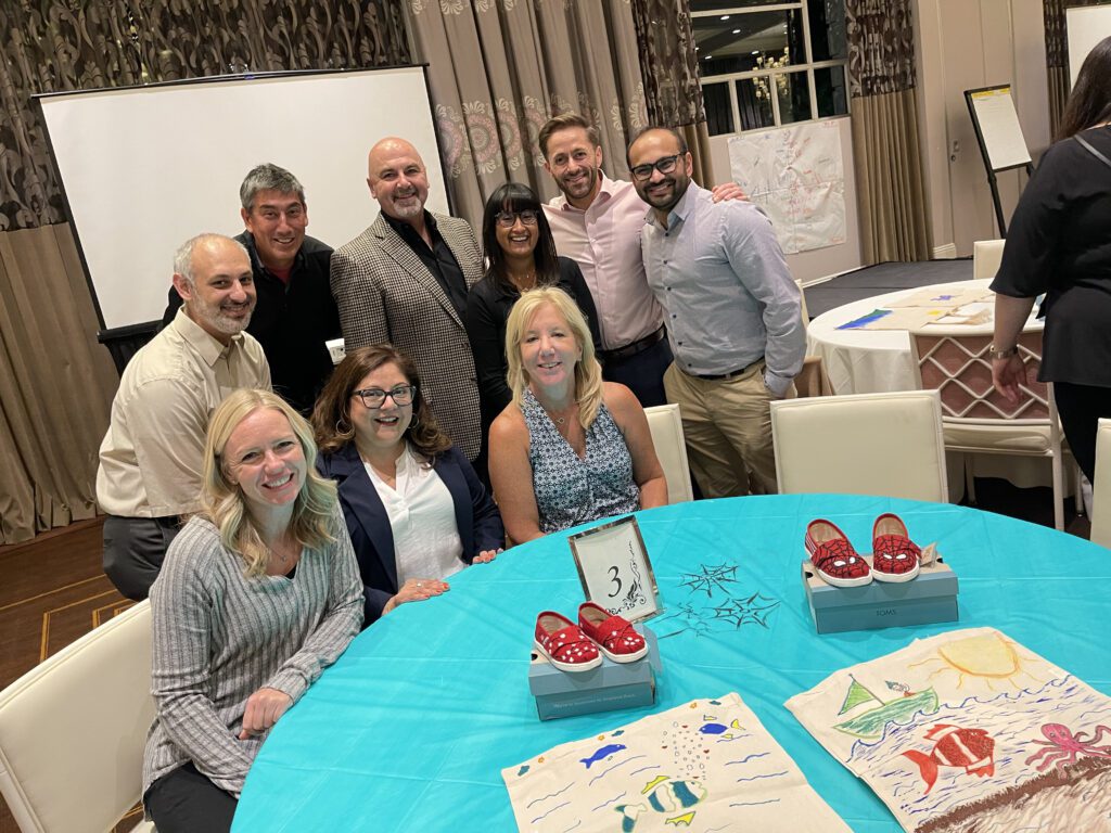 A group of participants smiles as they display their hand-painted TOMS shoes and tote bags during the Paint Soles for Souls charitable team building event. The red shoes and creatively designed bags will be donated to children in South America.