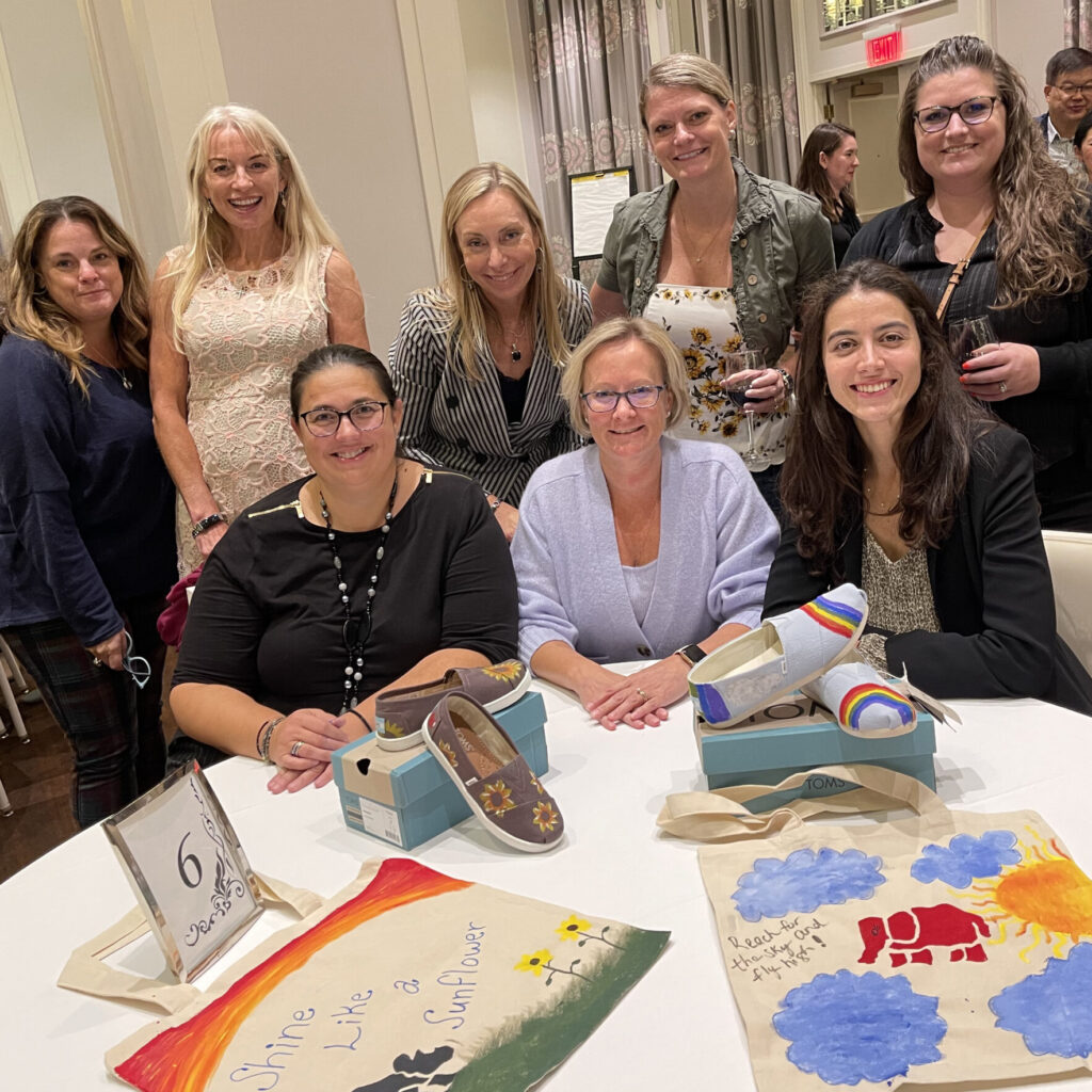 A group of participants smiles as they showcase their hand-painted TOMS shoes and tote bags during the Paint Soles for Souls charitable team building event. The colorful designs, including sunflowers and rainbows, are ready to be donated.