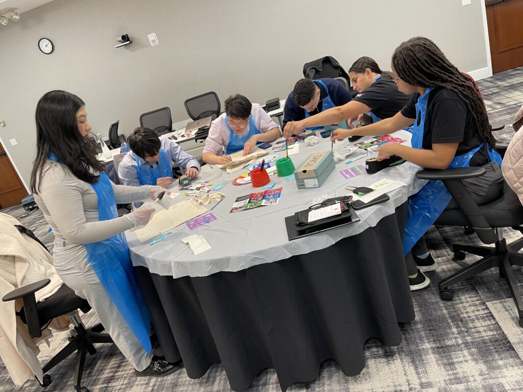 A group of participants wearing blue aprons sit around a table decorating TOMS shoes and tote bags during the Paint Soles for Souls charitable team building event. The team is fully engaged in the creative process, using paints and markers to design shoes that will be donated to children in South America.