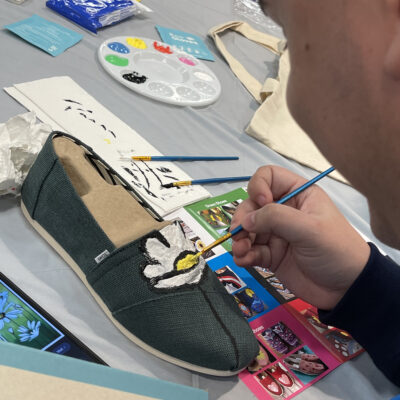 A participant carefully paints a white flower on a pair of green TOMS shoes during the Paint Soles for Souls charitable team building event. The table is covered with art supplies, including paintbrushes, a palette, and design inspiration cards.