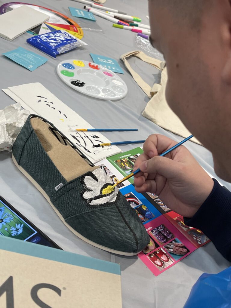 A participant carefully paints a white flower on a pair of green TOMS shoes during the Paint Soles for Souls charitable team building event. The table is covered with art supplies, including paintbrushes, a palette, and design inspiration cards.