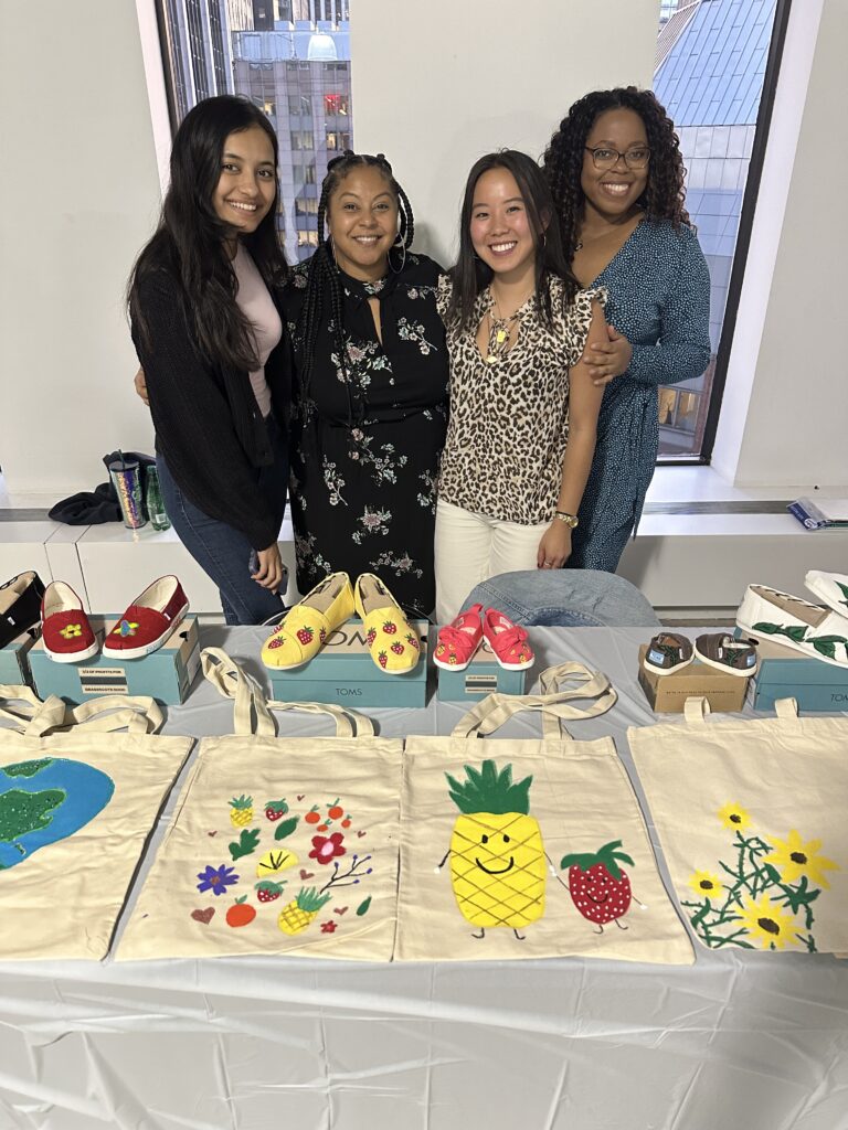 Four smiling participants stand behind a display of hand-painted TOMS shoes and decorated tote bags during the Paint Soles for Souls charitable team building event. The shoes and bags feature colorful designs, including flowers, pineapples, and strawberries, which will be donated to children in need in South America.