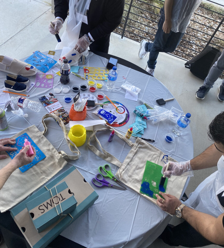 Participants decorate tote bags and TOMS shoes using stencils and paints during the Paint Soles for Souls charitable team building event. The table is covered with art supplies, including paintbrushes, scissors, and TOMS shoeboxes, as attendees creatively design items that will be donated.