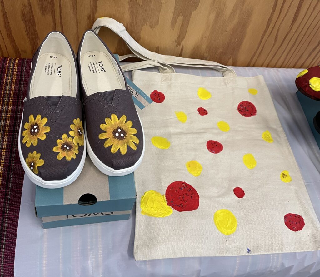 A pair of hand-painted TOMS shoes featuring sunflower designs, displayed alongside a tote bag decorated with yellow and red dots. These items were created during the Paint Soles for Souls charitable team building event and will be donated to children in South America.