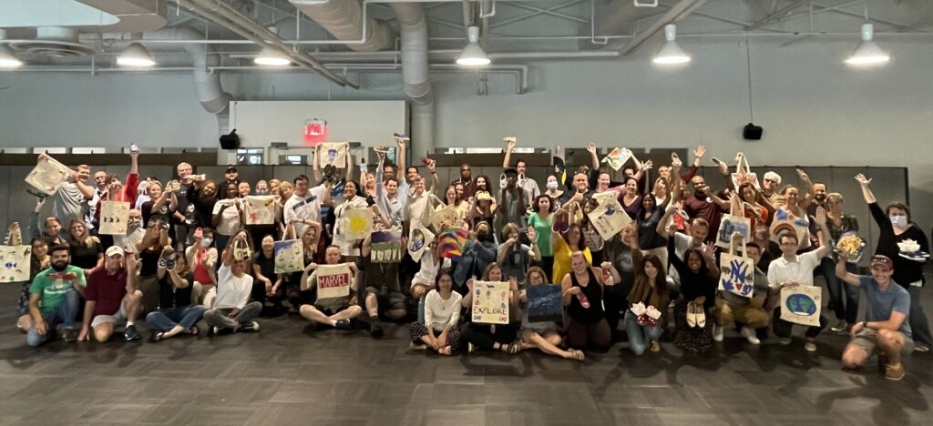 A large group of participants poses together, proudly holding up hand-painted TOMS shoes and tote bags they created during the Paint Soles for Souls charitable team building event. The group celebrates their creative contributions, which will be donated to children in South America.