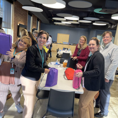 Group of smiling participants assembling colorful care packages for cancer patients during a TeamBonding event. The event, called Operation Cancer Care, combines fun team building activities with the meaningful goal of supporting cancer patients through the creation of care packages.