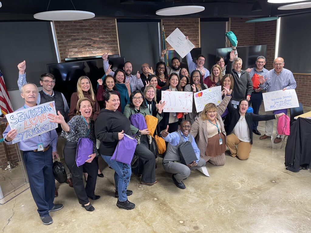 A large group of people posing together during a team building event. Some are holding colorful backpacks, while others hold hand-drawn supportive signs with messages like 'Sending Love & Strength.' The group is smiling, cheering, and making celebratory gestures, creating a positive and uplifting atmosphere.