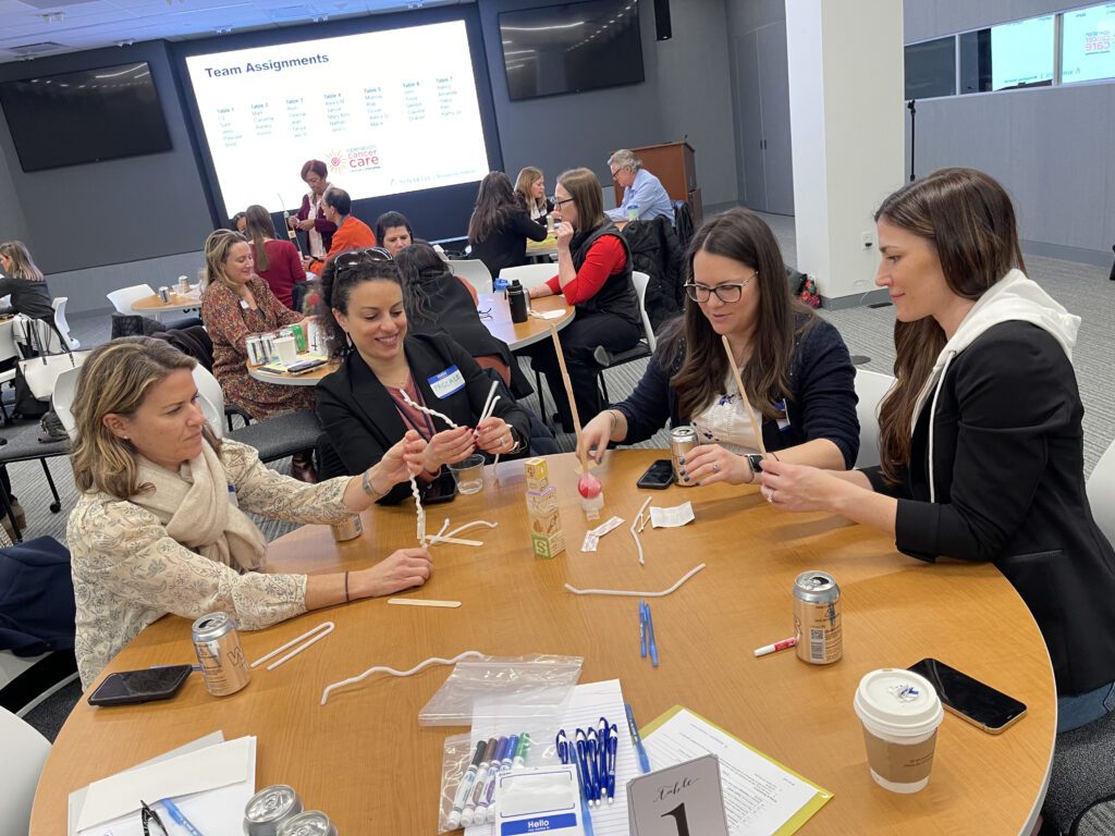 Four women are seated at a round table, engaging in a hands-on team building activity. They are using pipe cleaners, blocks, and small materials to build a structure. The room is filled with other groups participating in similar activities. A large screen in the background displays 'Team Assignments' and the event name, 'Operation Cancer Care.' The atmosphere is collaborative and focused, with various materials like markers, pens, and drinks on the table.